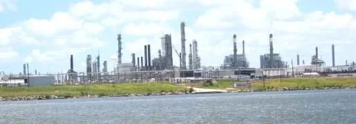 A photograph shows a view from the Houston Ship Channel towards a green shoreline. Just beyond the shoreline are numerous chemical towers with pipelines and roads connecting them.