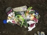Photo shows view from above of a hole in the ground filled with assorted trash (vegetable peelings, packaging wrappers, tissue, egg shells, tea bag, can lid, plastic lid).