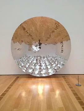 A photograph shows a sound mirror; a six-foot diameter concave disk fragmented by a repeating triangular pattern from a metal surface that distorts the reflection of its viewer into multiple images.