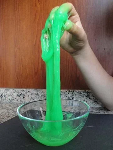A child’s hand stretches green slime upwards from a bowl.
