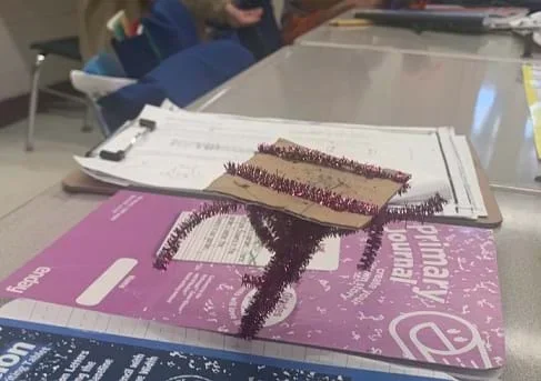 Image shows pink pipe cleaners holding up a piece of cardboard. Writing journals are placed on either side at the base of the creation.