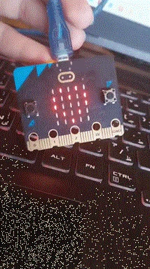 A photograph of a micro:bit connected to a computer with a USB cable and resting on a laptop keyboard. Several red LEDs in the micro:bit’s 5 × 5 LED display are illuminated in a pattern. The micro:bit’s two push buttons and five connection pins are also visible.