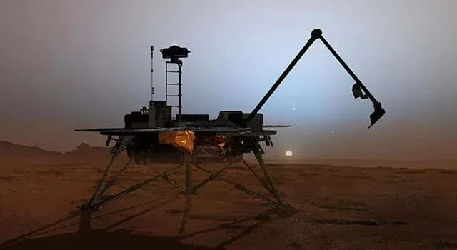 A picture showing an artist's rendering of the Phoenix Mars Lander sitting on the rust-colored Martian surface at twilight. The lander, supported by three metallic legs, has a distinctively long, thin robotic arm extended upward and to the right, ready for scooping or analysis. In the background, a hazy sky meets the horizon, with the faint disk of the sun or a moon visible just above the surface. The scene is dimly lit, casting deep shadows from the lander and its arm.
