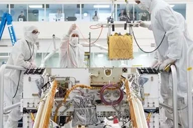 A photo showing three engineers wearing white protective cleanroom suits, gloves, and face masks working inside a laboratory to install a gold, cube-shaped instrument into the open body of a Mars rover. The rover is surrounded by exposed wiring, tubing, and mechanical components, with solar panels visible on either side.
