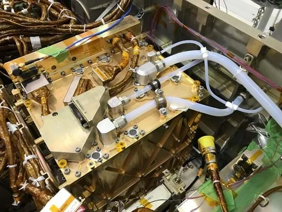 A photo showing a close-up view of the MOXIE instrument’s internal components, including gold-colored metal panels, silver fittings, and multiple translucent tubes connected to valves and ports.