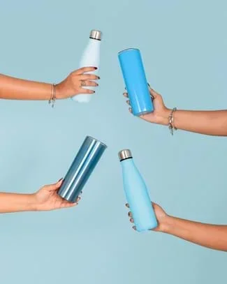 A photo showing four hands with diverse skin tones reaching in from the left and right sides of the frame against a solid pale blue background. Each hand holds a different reusable water bottle or tumbler in varying shades of blue.