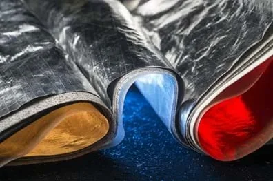A photo showing a close-up view of layered thermal insulation material. The outer layer is a crinkled, reflective silver foil. The textures appear metallic and industrial, set against a dark, speckled base.