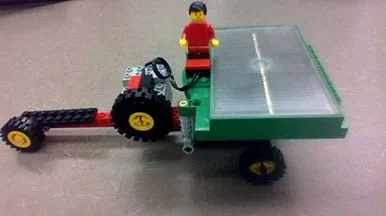 A photograph shows a small-size solar-powered wheeled vehicle made from an eLAB LEGO Renewable Energy Set. It has a long nose with a small wheel at the end and multiple wheels at the back, on which the solar panel is positioned. A LEGO person rides in the middle of the vehicle.