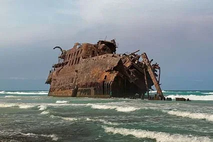 A large shipwreck is shown on choppy and wavy waters. The sky is blue and cloudy. The ship appears to be made out of metal, but is completely rusted with a brown/red color. The front of the ship is worn away and missing. One of the upper floors is angled and falling into the water.