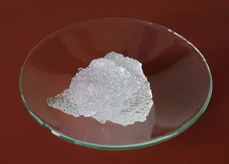 A photo showing a transparent glass petri dish placed on a reddish-brown surface. In the center of the dish, there is a pile of a foamy, white, porous material.