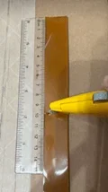A photo showing a close-up of a rectangular strip of orange-brown material placed next to a transparent plastic ruler. The ruler is marked in centimeters, with the material aligned along its length for measurement. A yellow box cutter is positioned at the edge of the material, with the blade extended, indicating that the material is in the process of being cut at 10 cm.