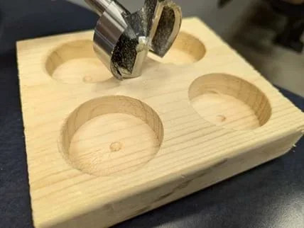 An image showing a close-up view of a Forstner bit resting on a wooden block. The wooden block measures approximately 6 inches by 2 inches by 6 inches. The block has four cylindrical holes drilled into it, each positioned in one of the four quadrants of the top surface. The holes are evenly spaced and appear smooth and well-defined, indicative of the clean cutting action of the Forstner bit. The Forstner bit itself, metallic and slightly worn from use, is positioned above one of the holes, laying on its side with the sharp edges visible.