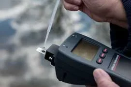 Photo shows a drop of liquid being placed on a sensor plate connected to a handheld plastic device with a small display screen and control buttons.