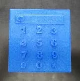 A photograph shows a thin blue square plastic piece that looks like a calculator. Raised on the surface is a short and wide rectangle with the numbers 1-9 below in three rows and the characters "@ 0 *" on the fourth row.