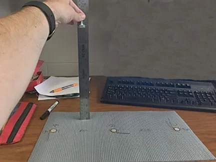 A photograph shows a person’s arm and hand holding a bolt 25 cm above a rubber mat (the same one shown/described in Figure 1) on a tabletop.