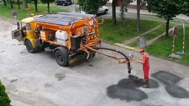 A photograph shows a road worker standing behind a truck in the middle of a street, using material flowing from a hose from the truck to patch potholes.