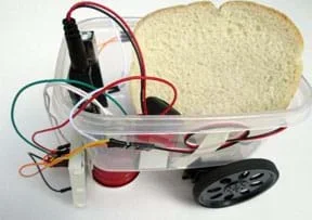 A photograph shows a clear plastic food container with two drive servomotors and their two attached wheels taped to one end (back) of the container, a small cup supporting the other end (front) of the container (the same bot frame as shown in Figure 2), plus sensors fastened to the front, a small breadboard taped to the outside front of the container, an Arduino microcontroller mounted inside front, and various wires that connect the servos, IR sensors and battery to the Arduino. It carries a slice of white bread.