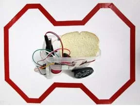 A composite photograph shows a continuous loop of red reflective tape on a 24 x 36-inch piece of white poster board. The route is a series of straight sections with periodic left and right turns, none greater than 45°. In the center is a Lunch-Bot carrying a slice of white bread. The Lunch-Bot is composed of a clear plastic food container with two drive servomotors with wheels taped to the bottom of one end of the container, a small cup taped to the bottom of the other end of the container (a support leg), sensors fastened to the front, a small breadboard taped to the outside front of the container, an Arduino microcontroller mounted inside front, and various wires that connect the servos, IR sensors and battery to the Arduino.