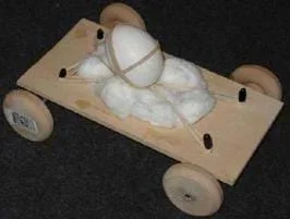 A photo shows a wooden platform with four wheels and an egg strapped with rubber bands to a foam layer on the platform.