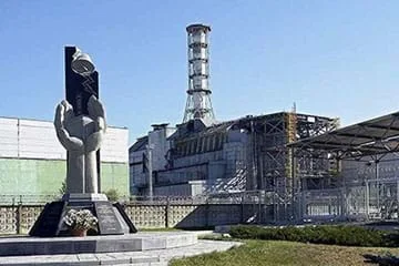 A photograph shows a monument shaped like a cupped pair of hands in front of the building remains of a power plant.