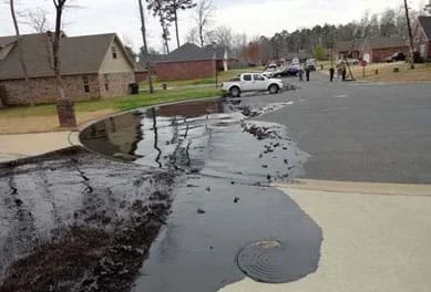 A photograph shows the black slick of an oil spill on a residential cul-de-sac street, spilling over a concrete driveway and contained in a pool by a curved concrete curb. The crude oil spill came from a rupture in ExxonMobil’s Pegasus 850-mile IL-to-TX pipeline in Mayflower, AR, in 2013.
