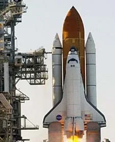 A photograph taken at Cape Canaveral, FL, shows a winged rocket launching next to a support structure. The rocket points upwards, has flames from its lower end, and is attached to three, un-winged fuel rockets (rocket boosters and external propellant tank), also pointed upwards.