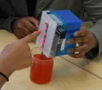 Photo of a hand holding a box with lighted LED bulbs, its probes inserted into a beaker of red liquid.