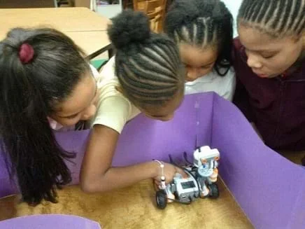 Students crowd over the construction paper walls of a model room containing a small wheeled robot.