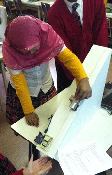 Photo shows a girl holding a small wheeled vehicle about midway on a ramp on a classroom desk.