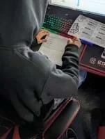 A photo showing a student wearing a dark hoodie seated at a computer station with a red-and-black esports-themed desk. The student is working on a printed worksheet titled "4 Input Logic Gate," using a pencil to complete a truth table, referencing a logic gates handout while looking at a screen that displays gate logic information.