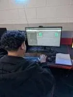 A photo of a student sitting at a computer desk using Wokwi, a logic circuit simulator, to test a custom logic gate design. The student is wearing a dark hoodie and is focused on the monitor, which displays logic gates, input switches, and a code window. A worksheet labeled “Logic Gates III” sits on the desk, partially completed with drawn gates and truth table outputs.