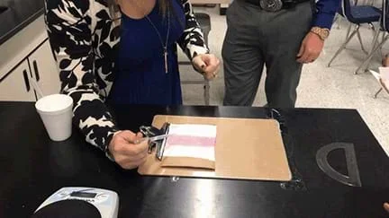 A woman’s arm with an eyedropper of liquid leans over a clipboard with a material showing a person testing the roll off angle.