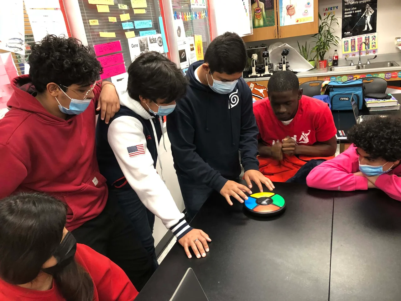 A photo of a student playing a tabletop version of “Simon” while five other students watch.