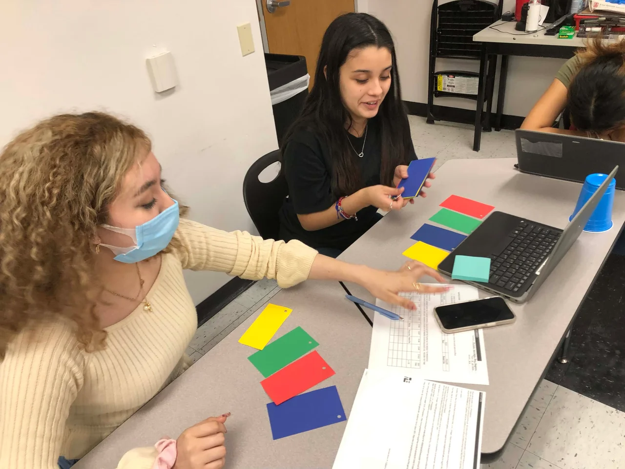 A photo of two students playing an analog “Simon” game with colored index cards; one is holding up a blue card and the other is about to start a timer.