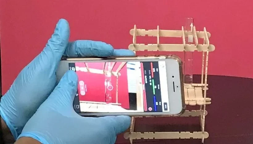 A student holds up a cell phone with a camera-based app open to measure the absorbance of food dye sample which is sitting in a sample holder.