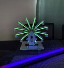 A small wooden model Ferris wheel sits on a table. The arms of the wheel are glowing a green color. A blacklight is on next to the Ferris wheel.