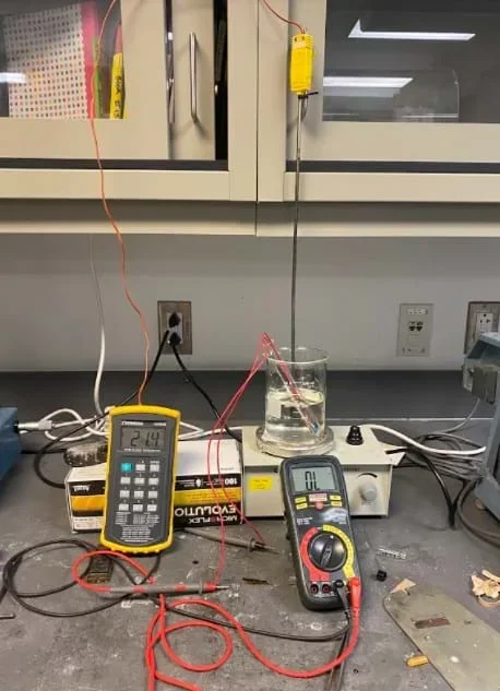 A photo showing the full set up for the resistance tests. A thermometer measures the temperature, a voltmeter measures the resistance, and a camera captures both screens to get the most accurate reading for the data collection.