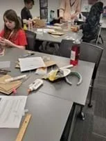 A photo of students gathering in groups at desks to work on prototypes using piping and classroom materials.