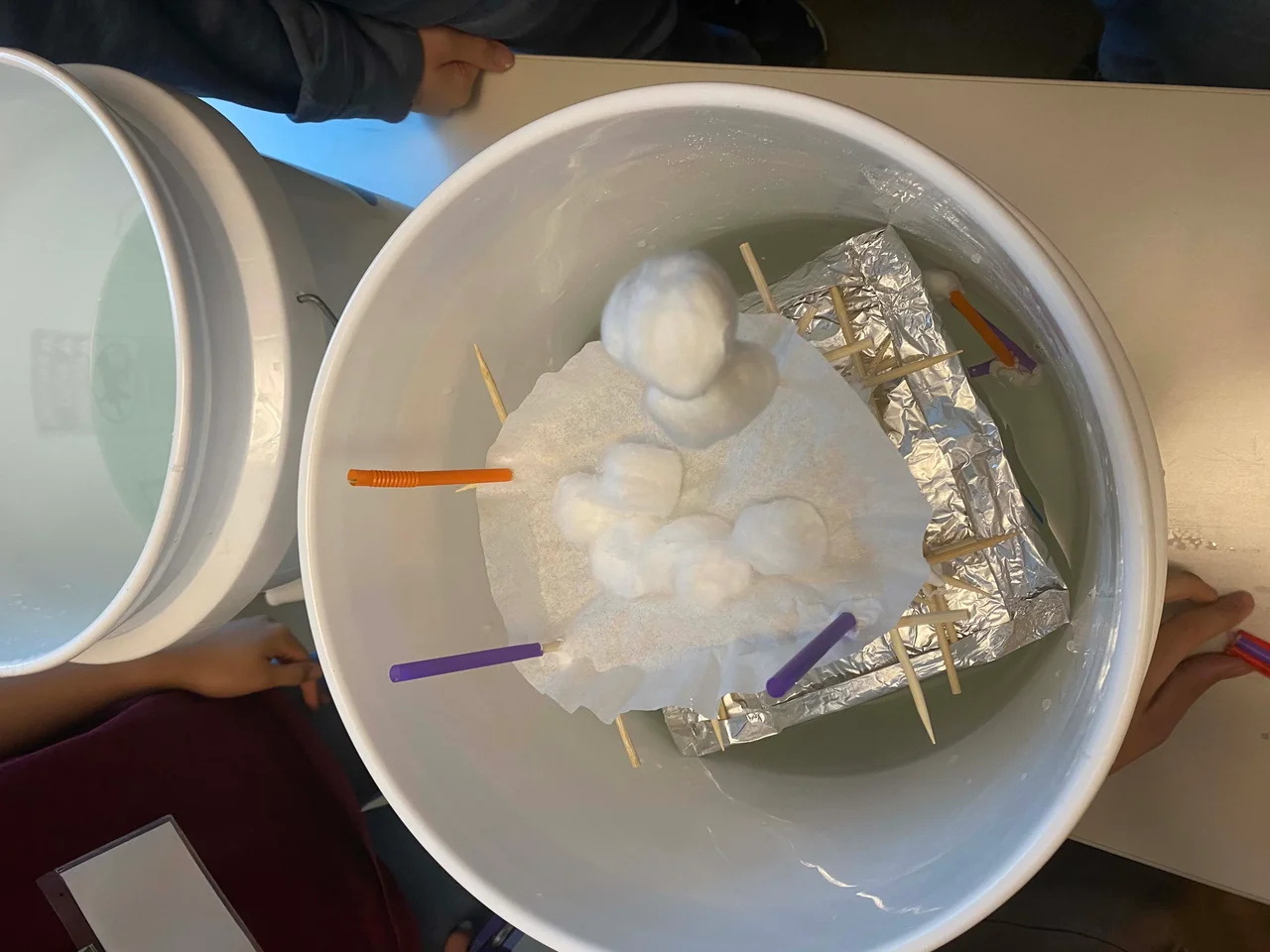 A white bucket is filled halfway with tap water; inside the bucket is a student group’s first prototype. The prototype is partially submerged in the water. Above the water line there is aluminum foil under some bamboo sticks that have a coffee filter full of cotton balls on top of it and some straw pieces branching off the top.