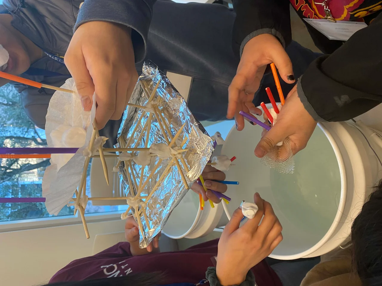 Three sets of student hands are placing an original prototype into a white bucket that is halfway filled with tap water. The prototype is held above the bucket of water. On the bottom of the prototype there is aluminum foil under some bamboo sticks which have a coffee filter full of cotton balls on top of it and some straw pieces branching off the top.