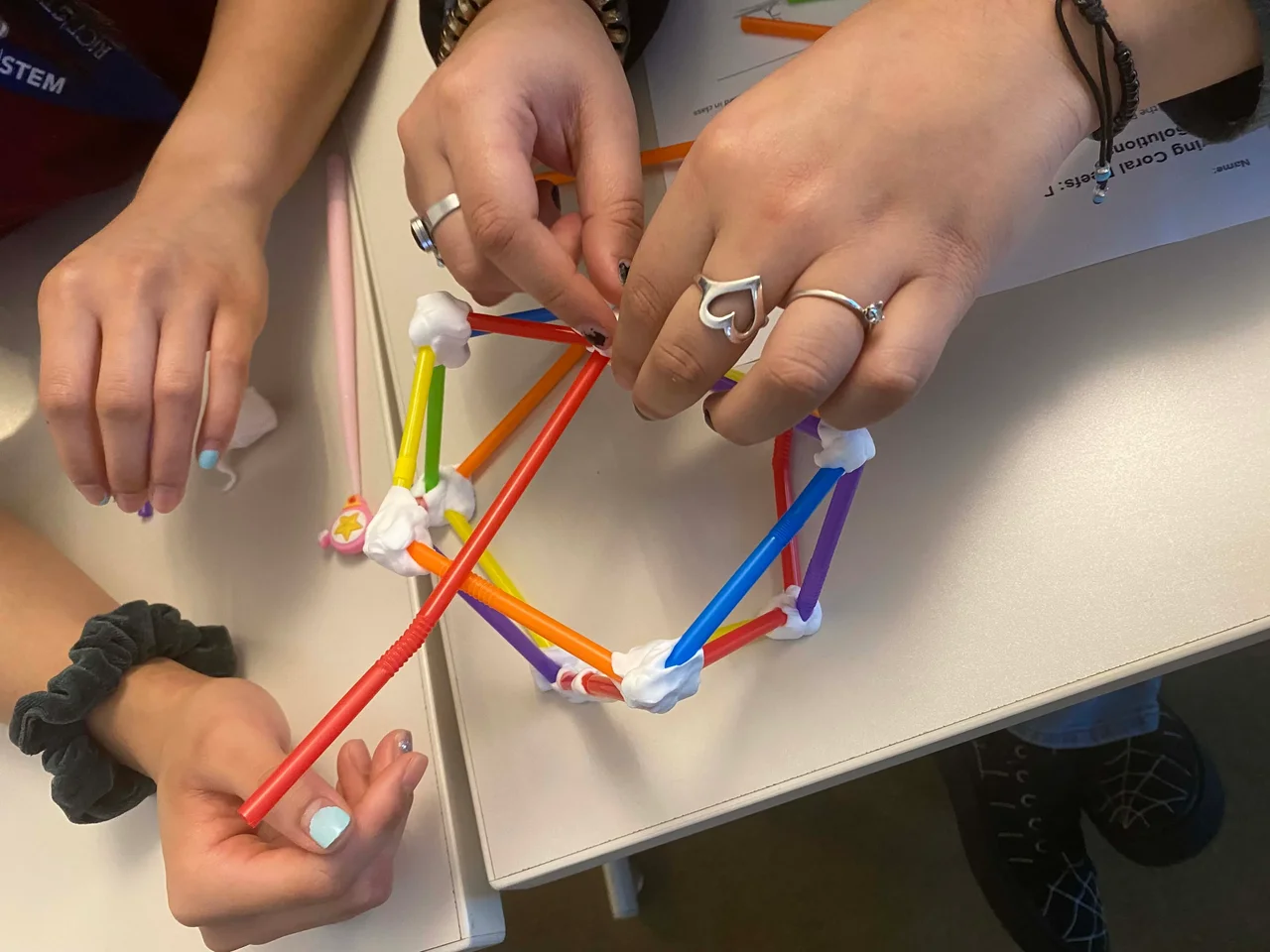Two sets of hands are shown above a desk putting together pieces of straw using putty to create a ball shape as part of the original design for their first prototype.