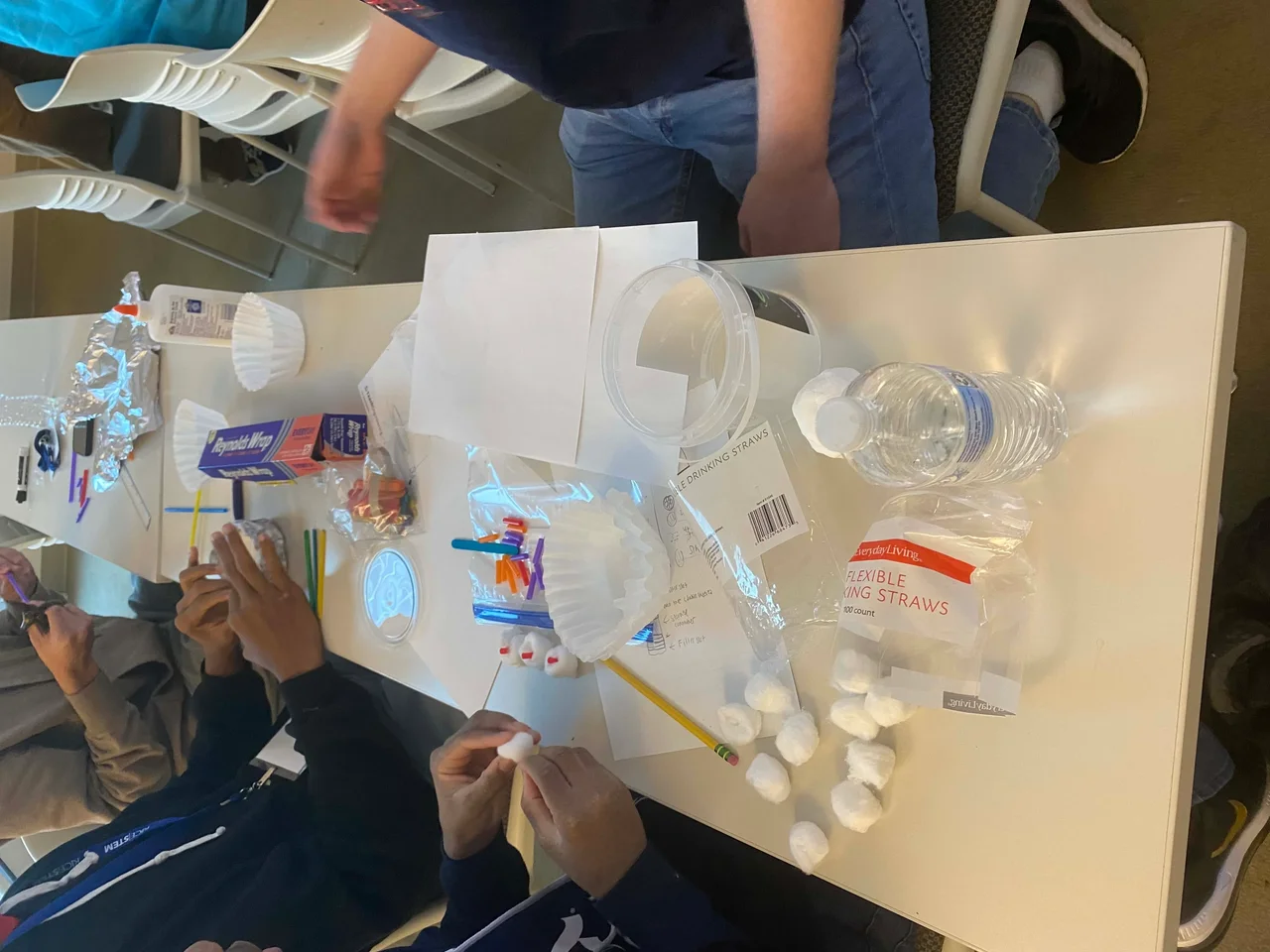 On a desk are several common household items such as aluminum foil, a coffee filter, cotton balls, pieces of paper, a water bottle, and some plastic trash. Two students (only hands included in the image) are starting to put together a prototype for their assignment.