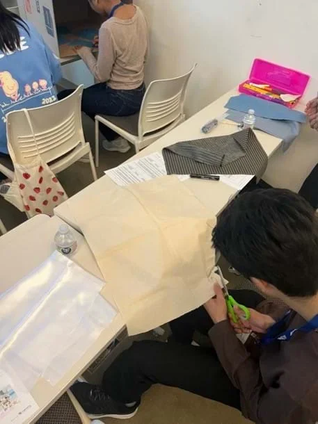 A photo of a student cutting a piece of beige cotton fabric using scissors.