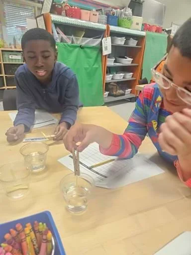 A photo showing one student holding a glucose testing strip grasped in a clothespin over a cup containing clear liquid while a second student looks on.