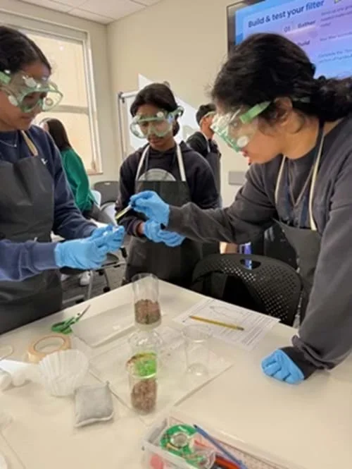 A photo showing three students, wearing protective gear, assembling the wet mount needed for viewing the microplastic solution under a microscope.