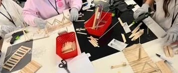 A photo showing three students at a worktable containing popsicles sticks, hot glue guns, syringe packets, plastic tubing, skewers, and other materials. They are constructing parts of their popsicle stick bridges.