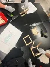 A photo showing two students constructing their bridges using popsicle sticks. In their work area, there is a basket of materials that includes popsicle sticks and glue sticks. They are using hot glue guns and protective gloves. Students also both have their bridge sketches for reference.
