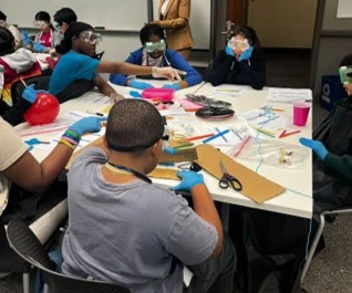 A photo showing students wearing safety goggles, aprons, and gloves collaborate at a table, using classroom materials to build a model.
