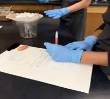 A photo showing two students wearing blue disposable gloves working at a black lab table. One student’s hands are visible in the foreground, holding a pencil above a sheet of white paper that has simple hand-drawn sketches and notes on it. An eraser rests on the paper. In the background, there is a clear plastic cup with a coffee filter placed inside it, forming a makeshift filtration setup. Another student is partially visible, with gloved hands positioned above the table. 