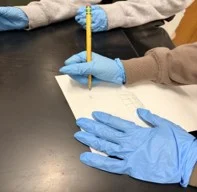 A photo showing two people seated at a black laboratory table. Both individuals are wearing disposable nitrile gloves. One person, wearing a brown long-sleeve shirt, is holding a yellow pencil and writing on a sheet of white paper. The paper contains a small hand-drawn sketch and a few short-written marks. Their gloved other hand rests flat on the paper. To the left, another person in a light gray sweatshirt has their gloved hands resting on the table, partially visible. The background includes part of a wood door and the edge of the table. 